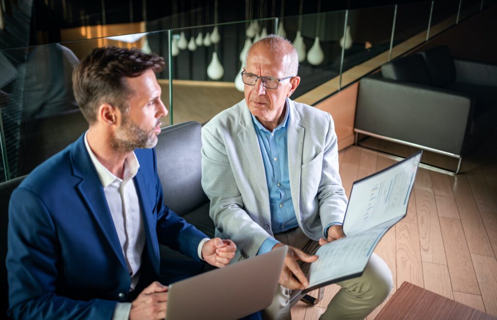 Businessman using laptop to discuss information with older colleague in modern business lounge