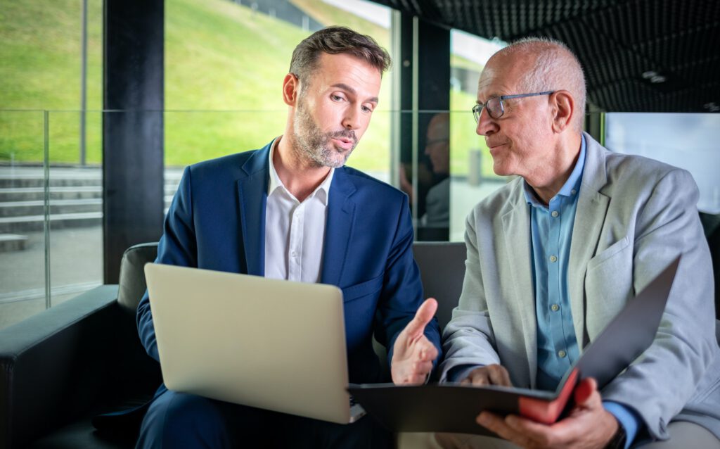 Businessman using laptop to discuss information with older colleague in modern business lounge