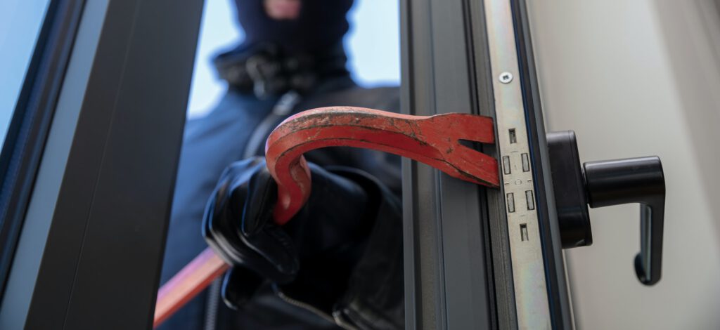 Thief in balaclava holding a crowbar to break a glass window.